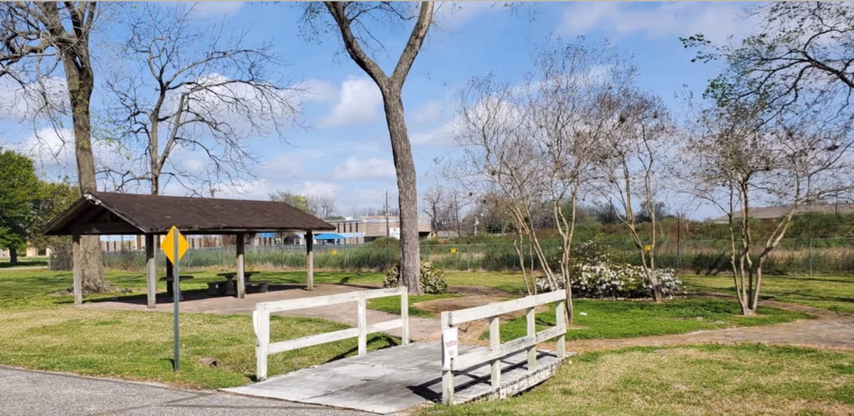 A grassy outdoor area with a small white wooden footbridge leading to a covered picnic pavilion surrounded by trees and shrubs under a blue sky.