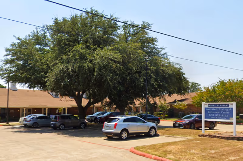 Parking lot and front of Mt Pleasant Assisted Living building with several cars, a large tree, and a freestanding facility sign.