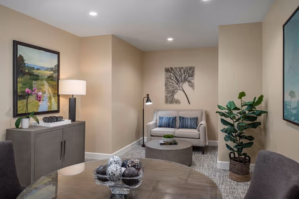A cozy living room area with a small beige loveseat adorned with two blue striped pillows, a round gray coffee table with books and a decorative bowl, a floor lamp, and a wall art piece resembling a tree branch. To the left, there is a gray cabinet with a table lamp, a potted orchid, and a framed landscape painting above it. A large green potted plant is placed on the right side of the room. In the foreground, part of a glass-top table with a decorative bowl filled with ornamental balls is visible.