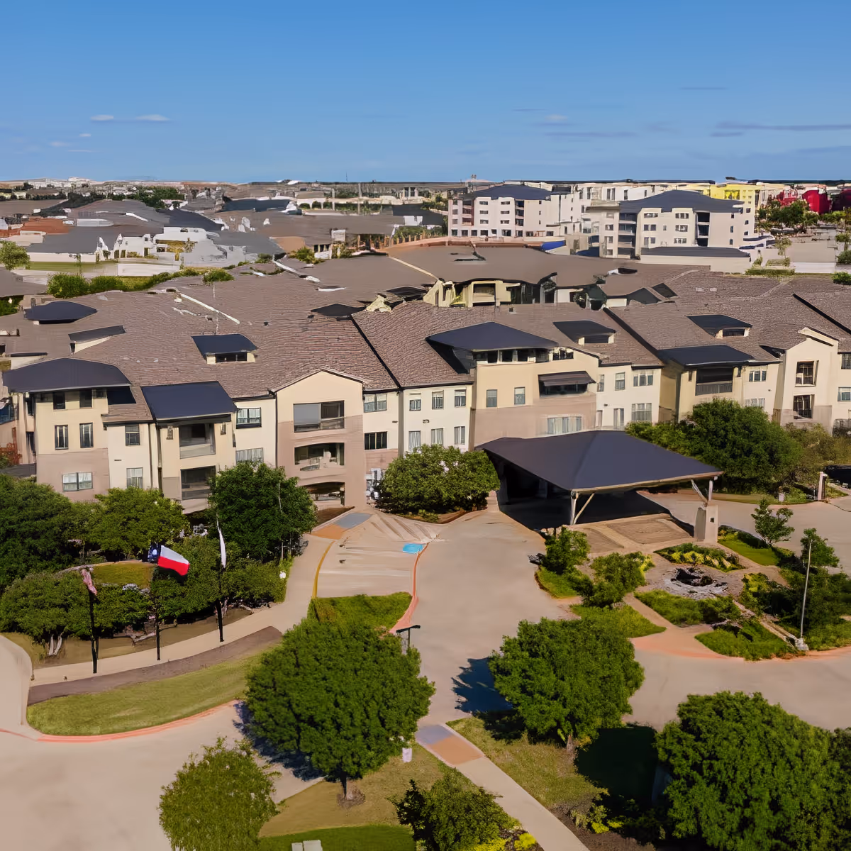 Aerial view of a large senior living facility with multiple connected beige buildings featuring dark roofs, surrounded by green trees and landscaped pathways under a clear blue sky.
