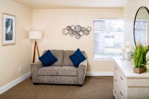 A cozy living room area with a gray loveseat adorned with two dark blue pillows. To the left of the loveseat is a tall floor lamp with a tripod base. Above the loveseat, there is a decorative wall art piece made of circular metal designs. To the right, a window with white blinds lets in natural light. A white dresser with a round mirror above it is positioned near the window, decorated with a green plant and candles.