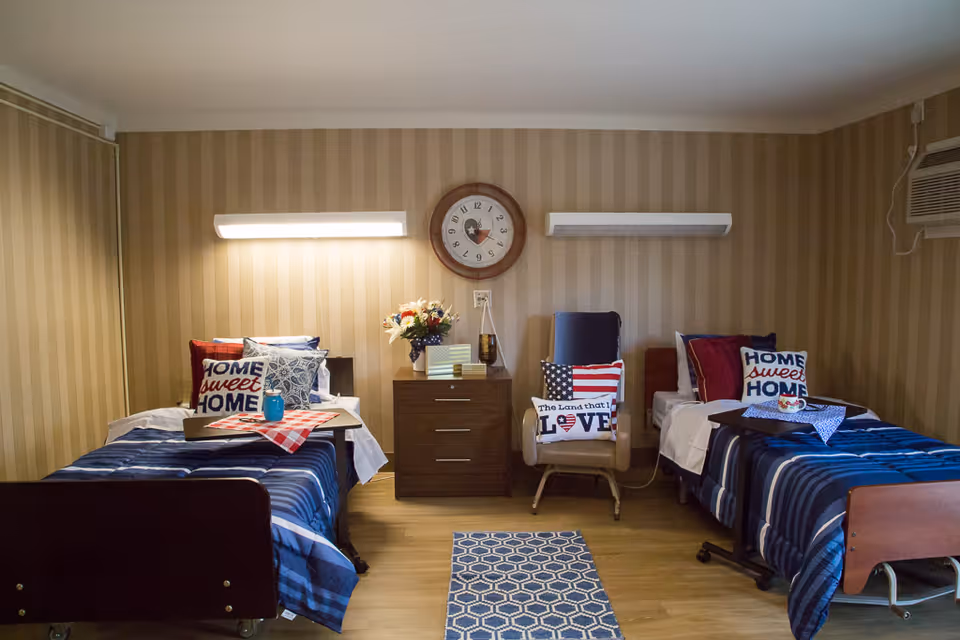 A room with two single beds, each with blue striped bedding and pillows that say 'Home Sweet Home'. Between the beds is a wooden nightstand with a vase of flowers, books, and a clock on the wall above. There is a chair with patriotic-themed pillows and a small blue patterned rug on the wooden floor.
