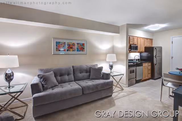 Interior view of a senior living facility apartment featuring a gray sofa with two matching pillows, two glass-top side tables each with a lamp, a colorful framed artwork on the wall, and a kitchen area with wooden cabinets, stainless steel appliances including a refrigerator, stove, and dishwasher. A small dining table with chairs is partially visible on the right.