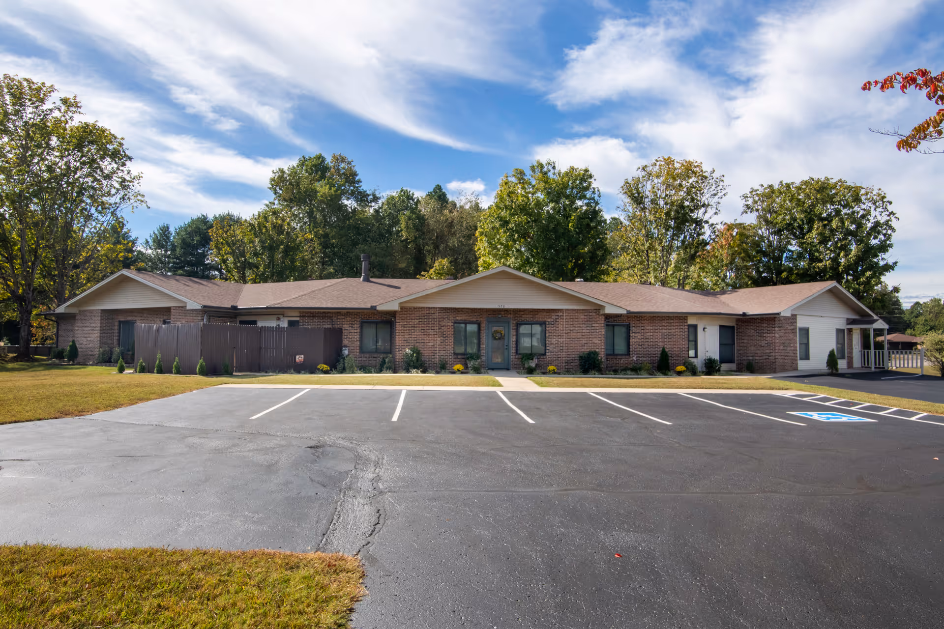 Single-story brick building with a brown roof surrounded by trees under a partly cloudy sky. There is a paved parking lot with marked parking spaces, including a handicapped spot, in front of the building.