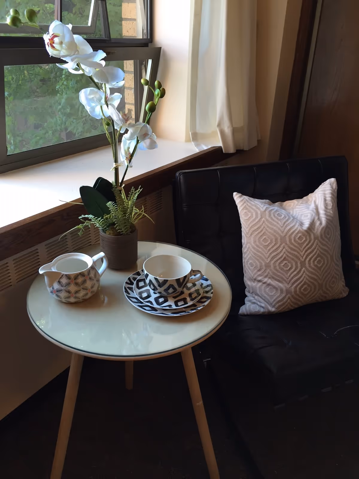 A small seating corner with a round side table holding a potted orchid, a teacup and creamer beside a black cushioned chair and window.