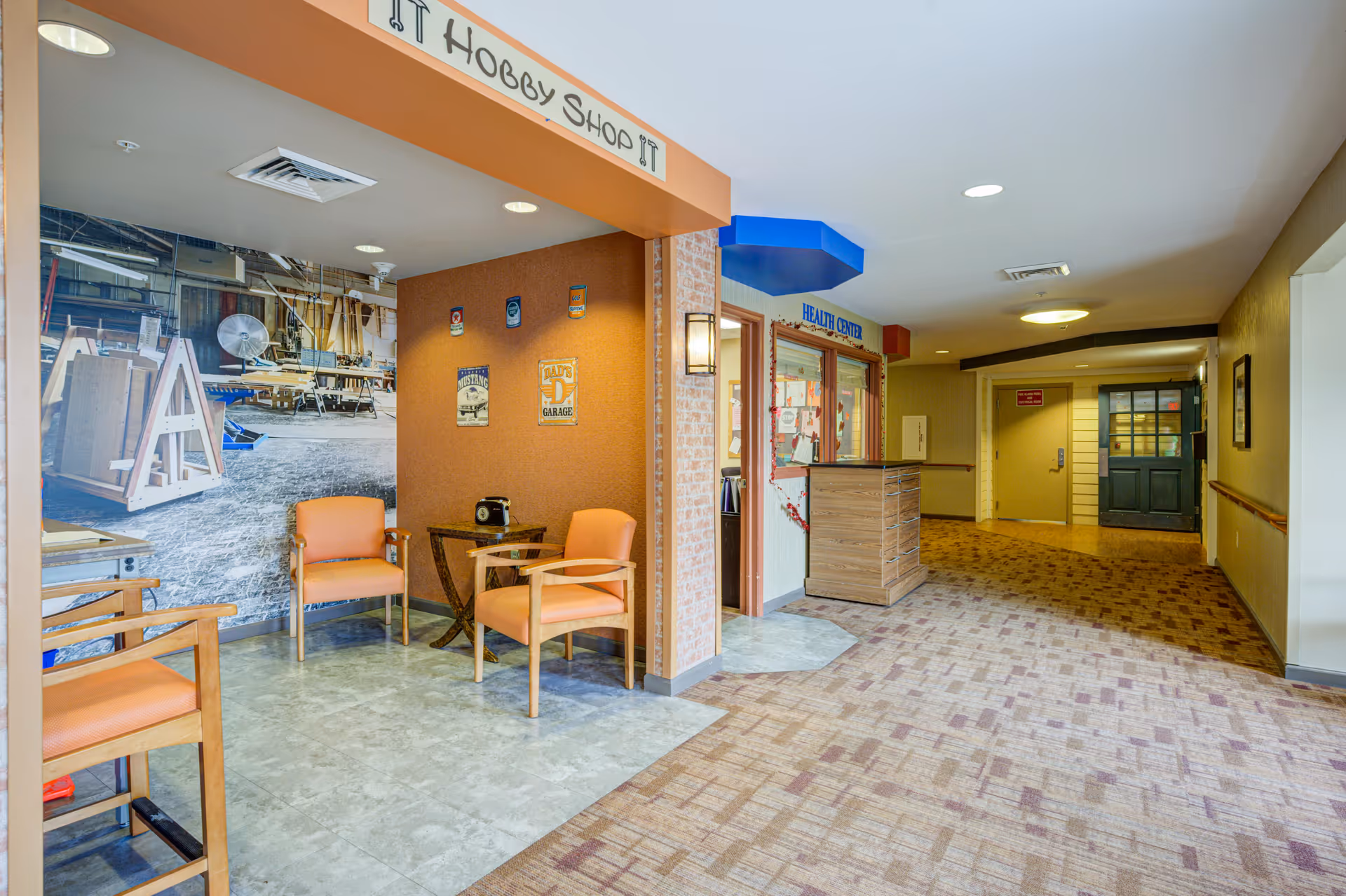 Interior hallway of a senior living facility with a small seating area labeled 'Hobby Shop' featuring three orange chairs and a small table. The hallway has carpeted flooring and leads to a health center with a wooden reception desk. The walls are decorated with signs and a large mural of a workshop.