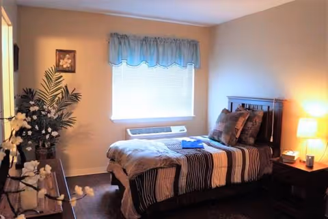 A neatly made bedroom with bed, nightstand, lamp, and window in soft lighting.