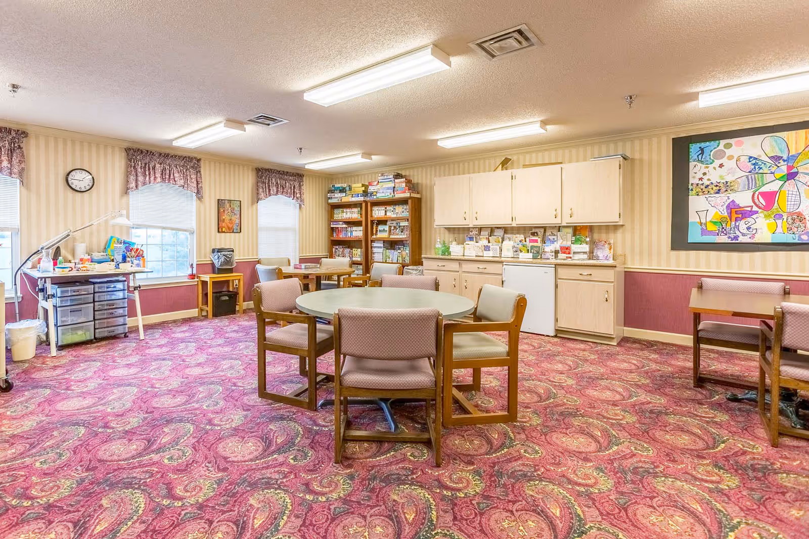 Bright senior community activity room with round table and chairs, cabinets, bookshelves, and craft supplies.