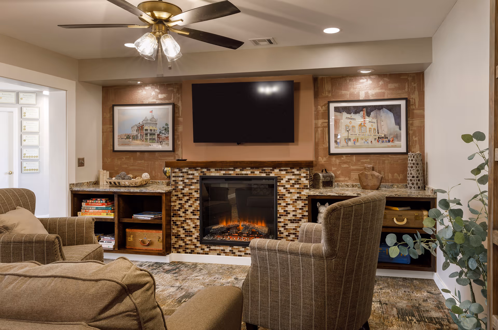 Cozy living room with armchairs facing a tiled fireplace and wall-mounted TV, built-in shelving, artwork, and a ceiling fan.