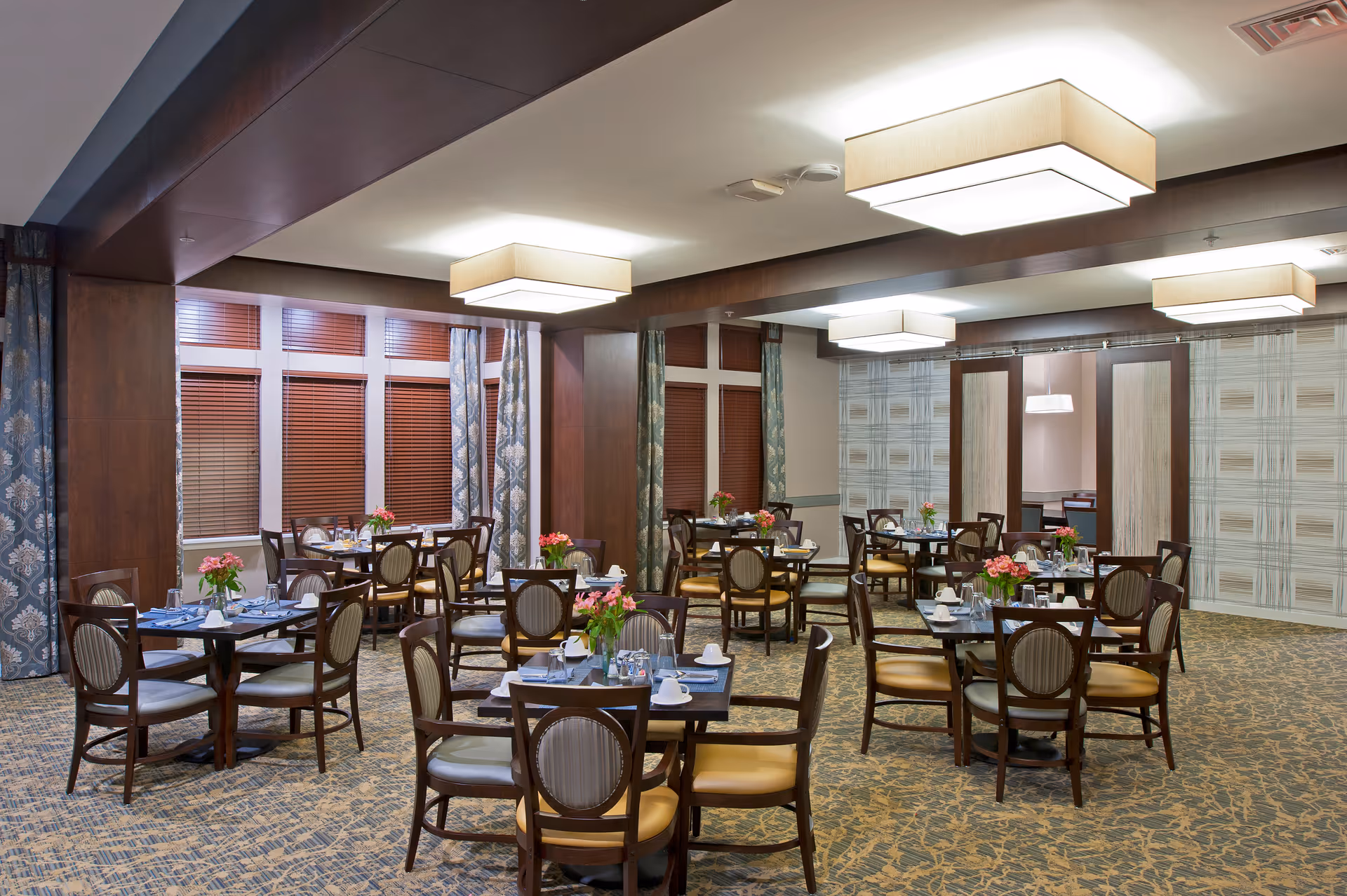 A well-lit dining room with multiple tables set for meals. Each table has chairs around it, place settings with cups, plates, and silverware, and a small vase with pink flowers. The room features large windows with wooden blinds and patterned curtains, carpeted floor, and modern rectangular ceiling lights.