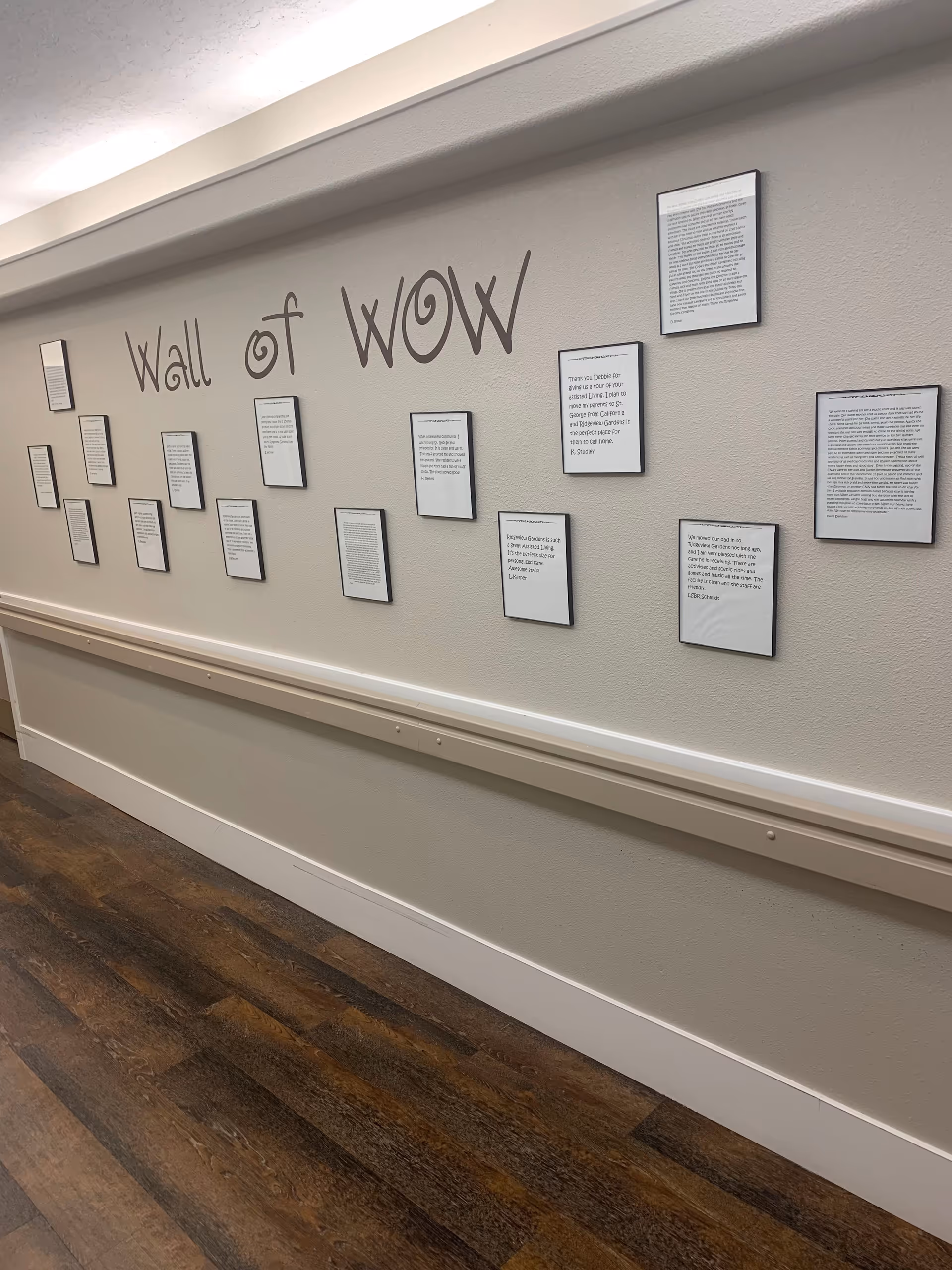 A hallway wall display titled 'Wall of WOW' featuring multiple framed notes or letters arranged in a row on a light-colored wall above a handrail, with wood flooring beneath.