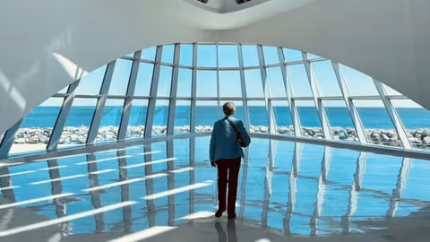 A person stands in a bright, modern interior with large floor-to-ceiling windows looking out over the ocean and sunlight reflecting on the shiny floor.