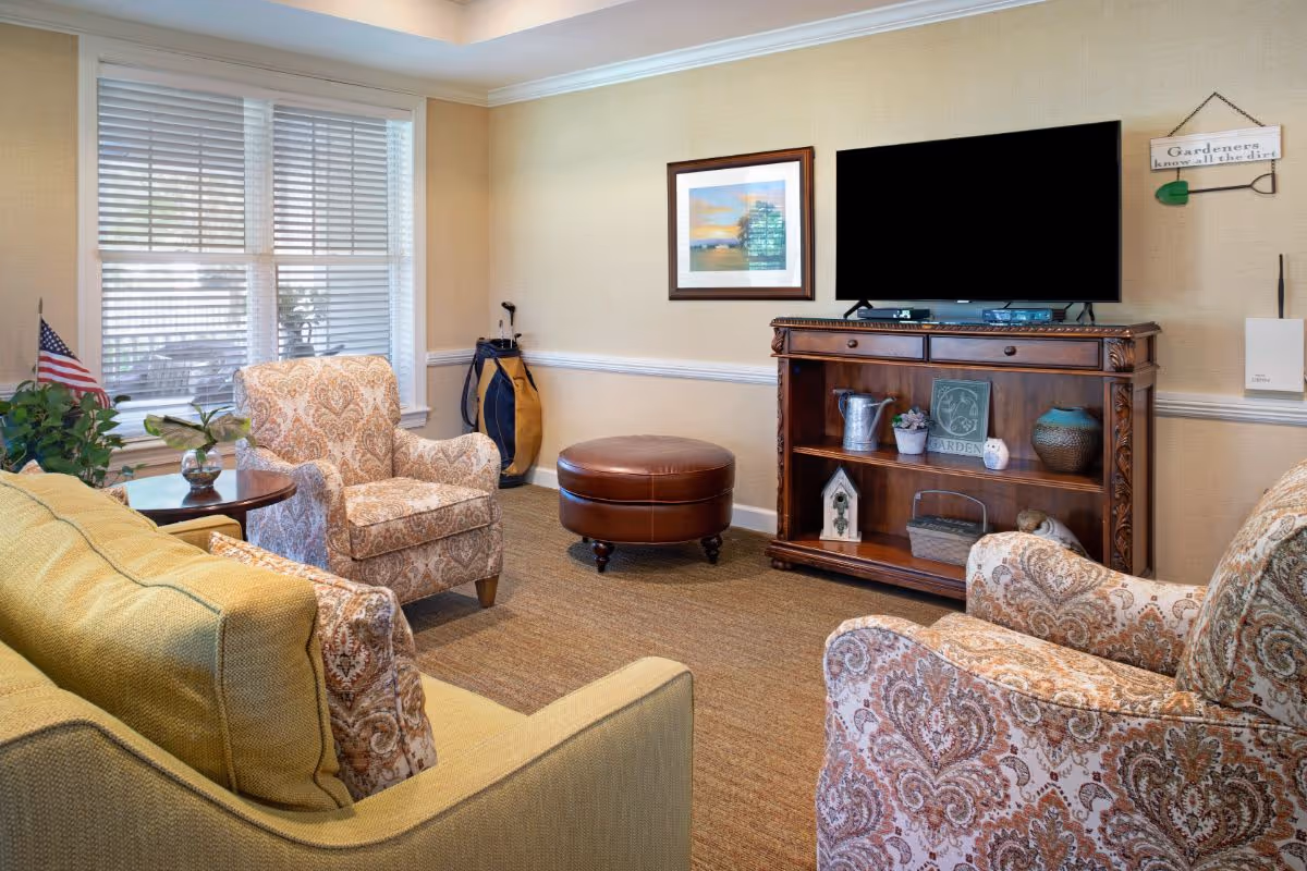 A cozy living room with a yellow sofa, two patterned armchairs, a round brown leather ottoman, a wooden TV stand with decorative items, a flat-screen TV mounted on the wall, a window with white blinds, and a small side table with a plant and an American flag.