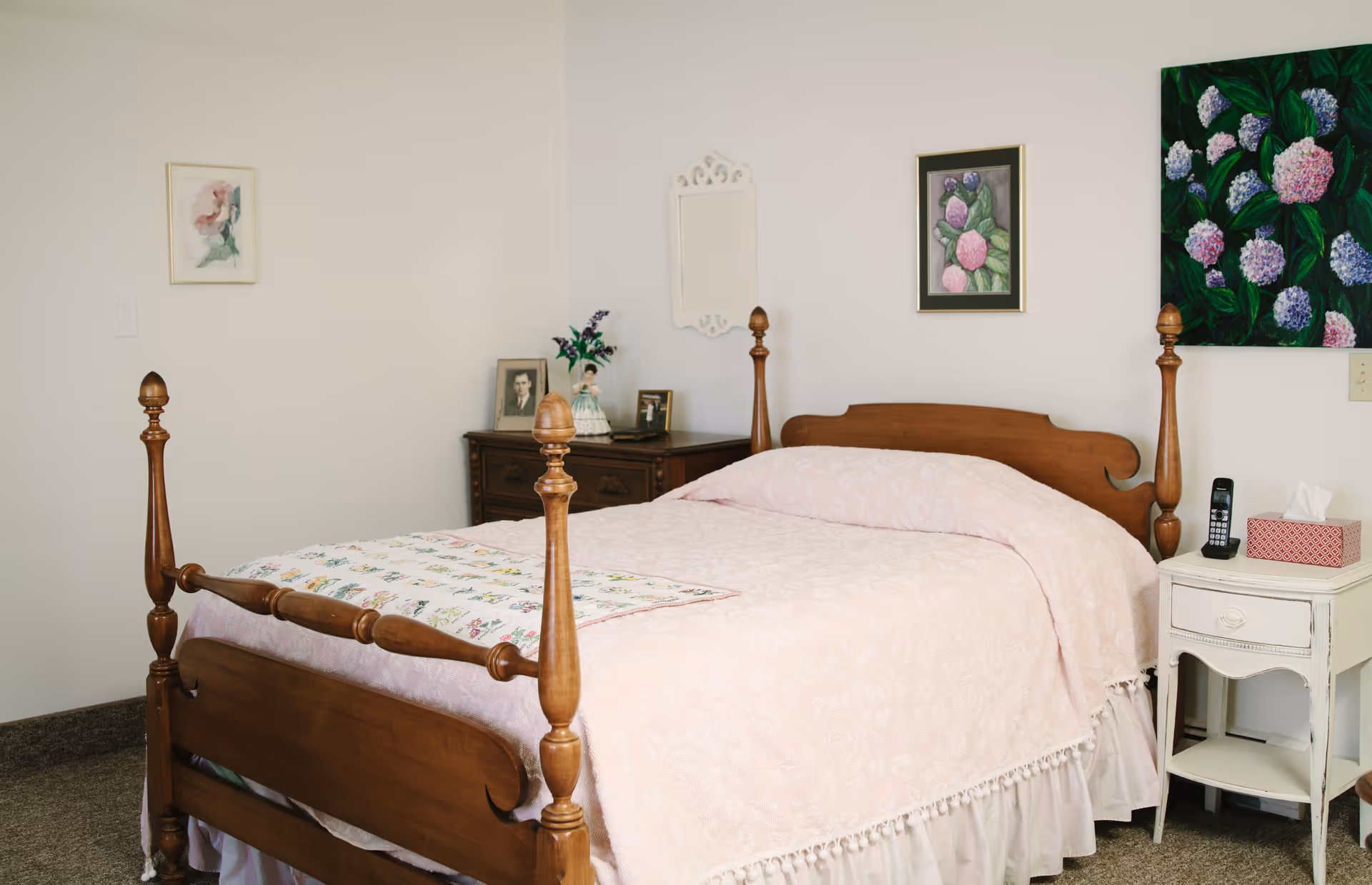 A cozy bedroom with a wooden four-poster bed covered in a light pink bedspread. There is a white nightstand with a cordless phone and a tissue box on it. On the wall above the nightstand are two framed floral paintings. A small dresser with framed photos and a vase with flowers is in the corner. The walls are light-colored and the carpet is brown.