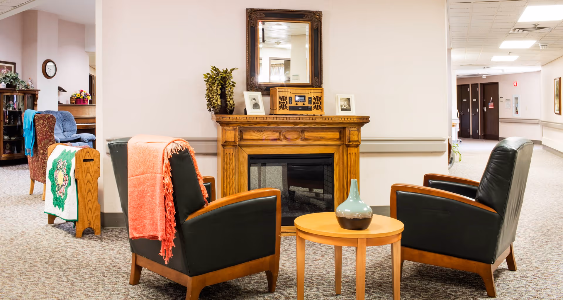 A cozy seating area in a senior living facility with two black leather armchairs draped with colorful throws facing a wooden electric fireplace. A small round wooden table with a decorative vase is placed between the chairs. In the background, there is a hallway and additional seating with a blue armchair and a wooden magazine rack.