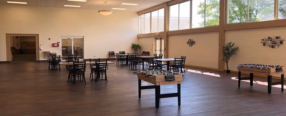A spacious room with wooden flooring featuring several tables and chairs arranged for seating. Two foosball tables are positioned in the foreground. Large windows near the ceiling allow natural light to fill the room. The walls are decorated with minimalistic metal wall art and there are a couple of potted plants. The room appears to be a common area or lounge space.