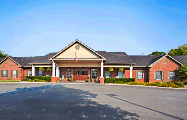Front exterior view of a single-story brick building with a covered entrance supported by white columns. The building has multiple windows with green shutters and hanging plants near the entrance. An American flag is displayed above the entrance. The sky is clear and blue.