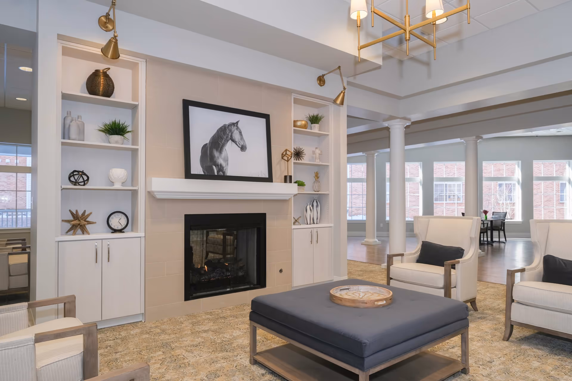 A bright and spacious living room area with a fireplace in the center, flanked by built-in white shelves decorated with vases, plants, and decorative objects. Above the fireplace is a black and white framed photo of a horse. The room features beige walls, a patterned carpet, two beige armchairs with dark cushions, and a large dark ottoman with a wooden tray on top. In the background, there are large windows letting in natural light and white columns supporting the ceiling.