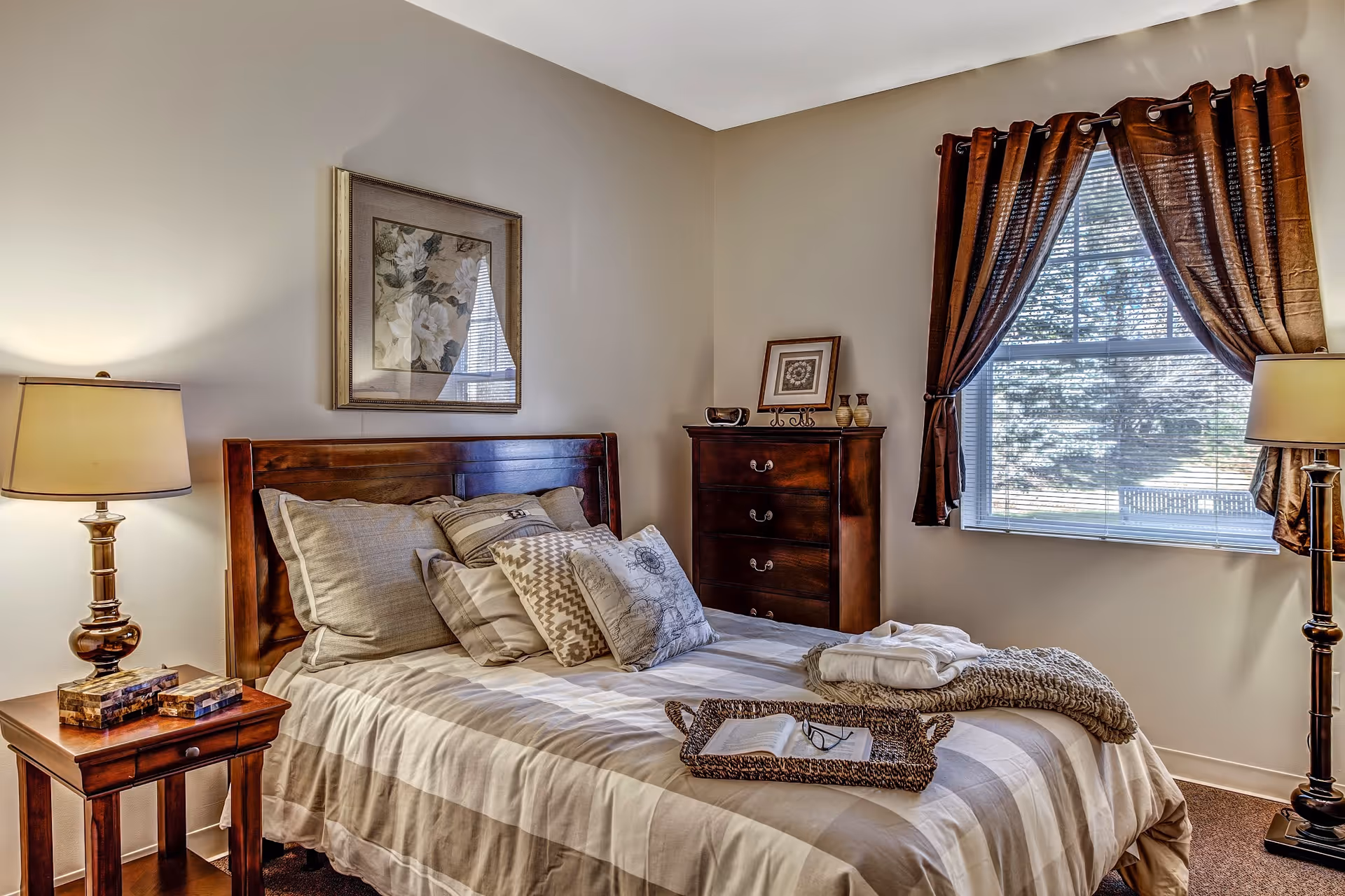 A cozy bedroom with a wooden bed frame, neatly made bed with multiple pillows, a bedside table with a lamp and books, a wooden dresser with decorative items, and a window with brown curtains letting in natural light.