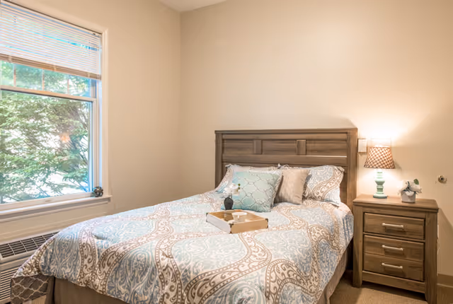 A cozy bedroom with a wooden bed frame and a neatly made bed featuring patterned bedding in light blue and beige tones. There are three pillows on the bed, and a wooden tray with a small decorative item is placed on the bedspread. Next to the bed is a wooden nightstand with three drawers, a small lamp with a patterned lampshade, and a small potted plant. A window with blinds partially open shows greenery outside.