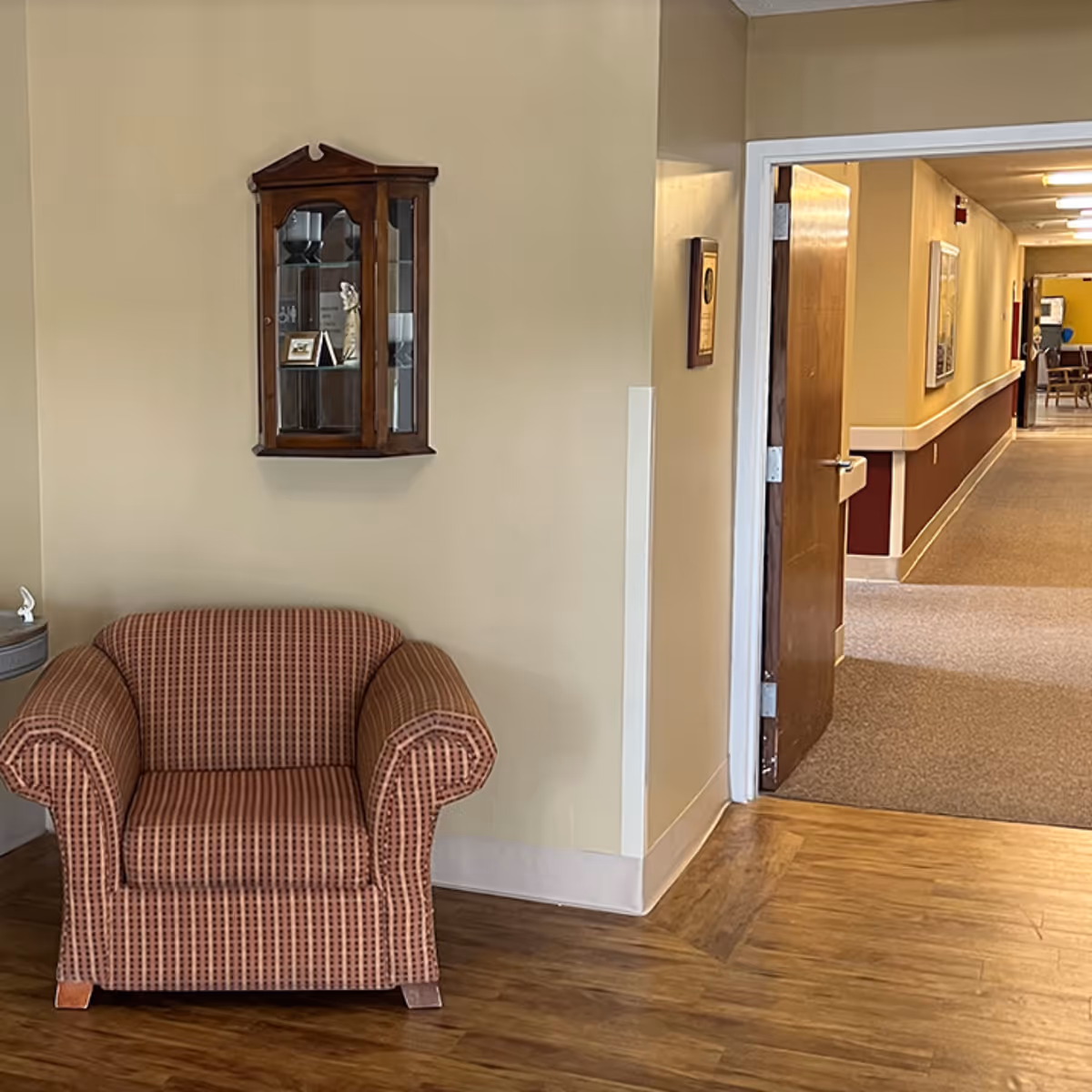 A patterned armchair beside a beige wall and a wooden display cabinet in a hallway.