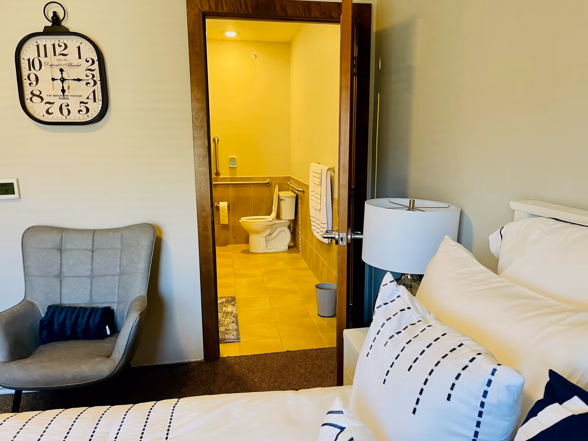 View of a bedroom with a bed featuring white and navy blue pillows and bedding, a white nightstand with a lamp, a gray armchair with a dark blue cushion, and a wall clock. The door to an adjacent bathroom is open, showing a toilet, towel rack with white towels, grab bars, and tiled floor and walls.