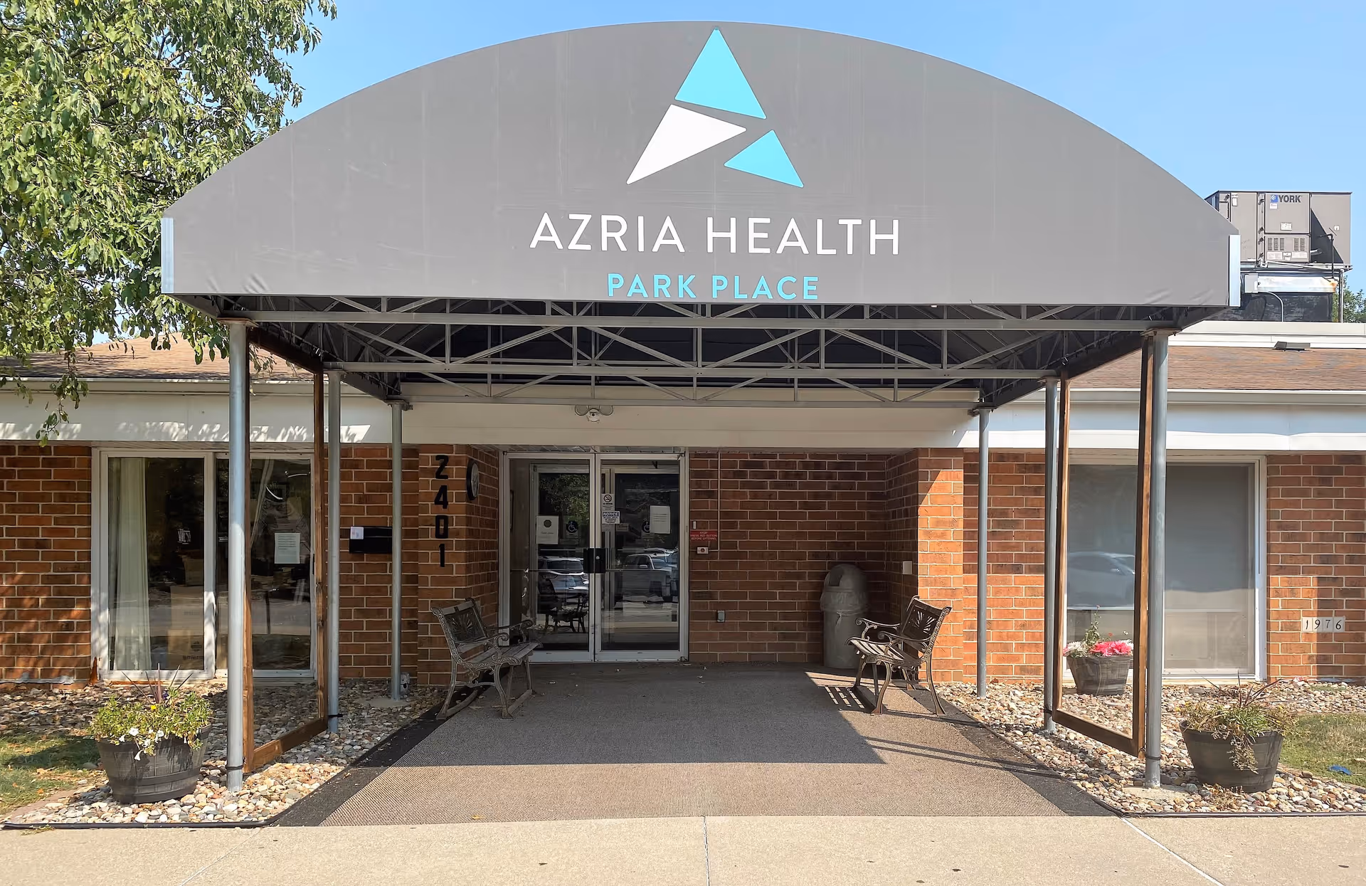 Entrance of Azria Health Park Place facility with a covered walkway, two benches on either side, brick walls, glass double doors, and potted plants near the entrance.