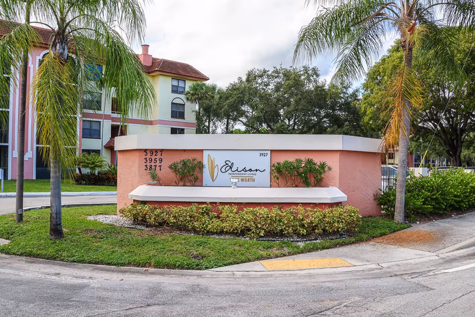 Exterior view of the entrance sign for Elison Independent Living of Lake Worth, surrounded by palm trees, shrubs, and a curved driveway with a multi-story building in the background.