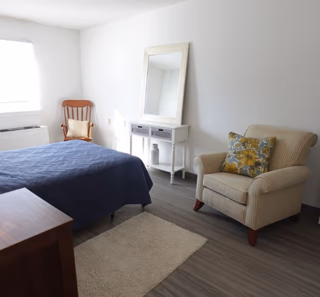 A bright bedroom with a navy-covered bed, a beige armchair with a floral pillow, a white console with a mirror, and a wooden rocking chair by the window.