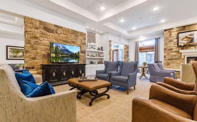 A cozy living room area in a senior living facility featuring a stone accent wall with a mounted flat-screen TV displaying a nature scene. The room includes various seating options such as a beige sofa with blue pillows, two patterned armchairs, and two brown leather chairs. There is a wooden coffee table in the center and a fireplace on the right side with a framed picture above it. The room is well-lit with recessed ceiling lights and has large windows with curtains allowing natural light to enter.