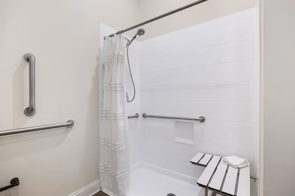 A clean, accessible shower area with white tiled walls, a white shower curtain, a handheld showerhead, and stainless steel grab bars on the walls. There is a foldable white bench with a folded towel on it inside the shower.