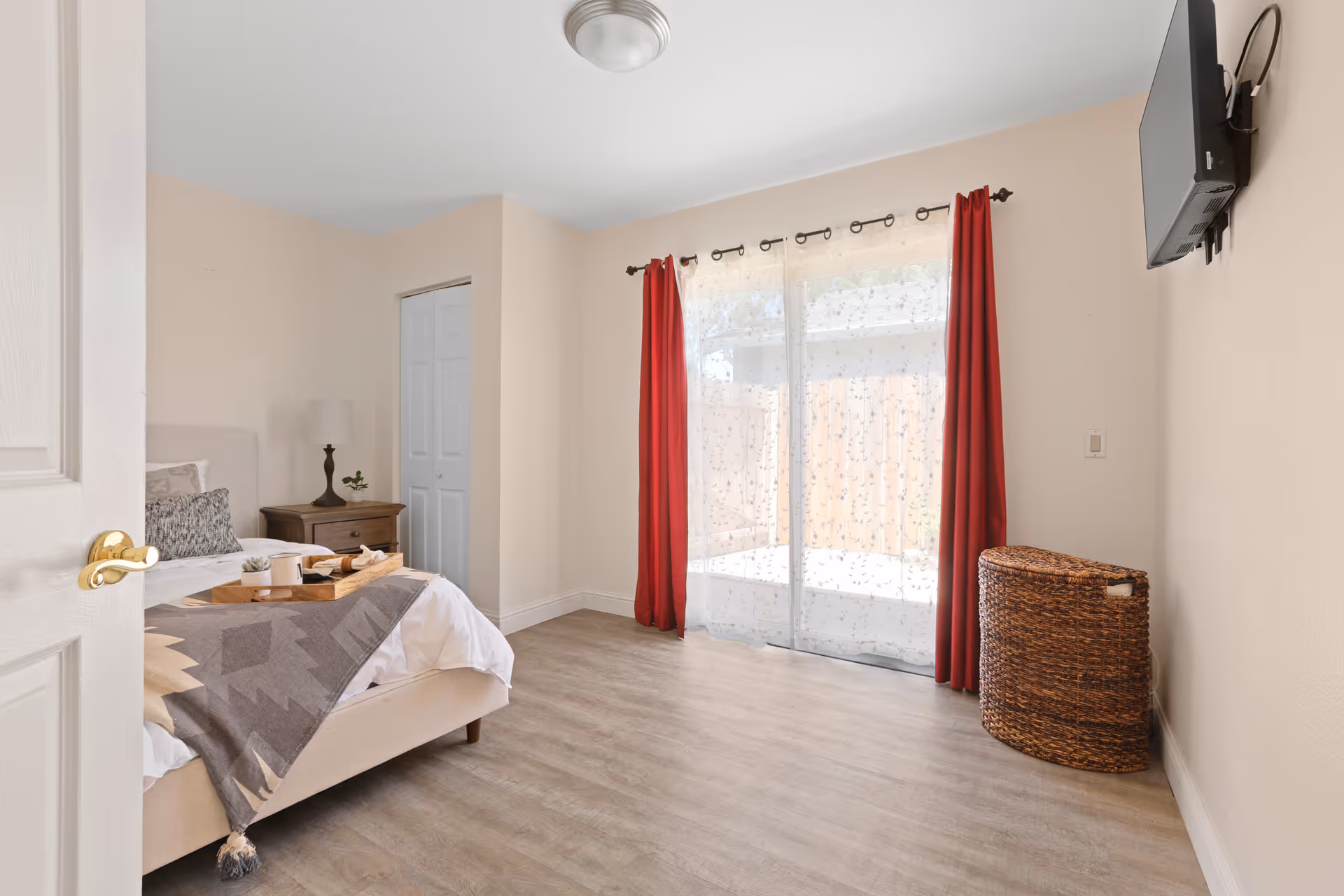 Bright bedroom with a bed and nightstand, sliding glass doors with red curtains, a wall-mounted TV, and a wicker hamper.