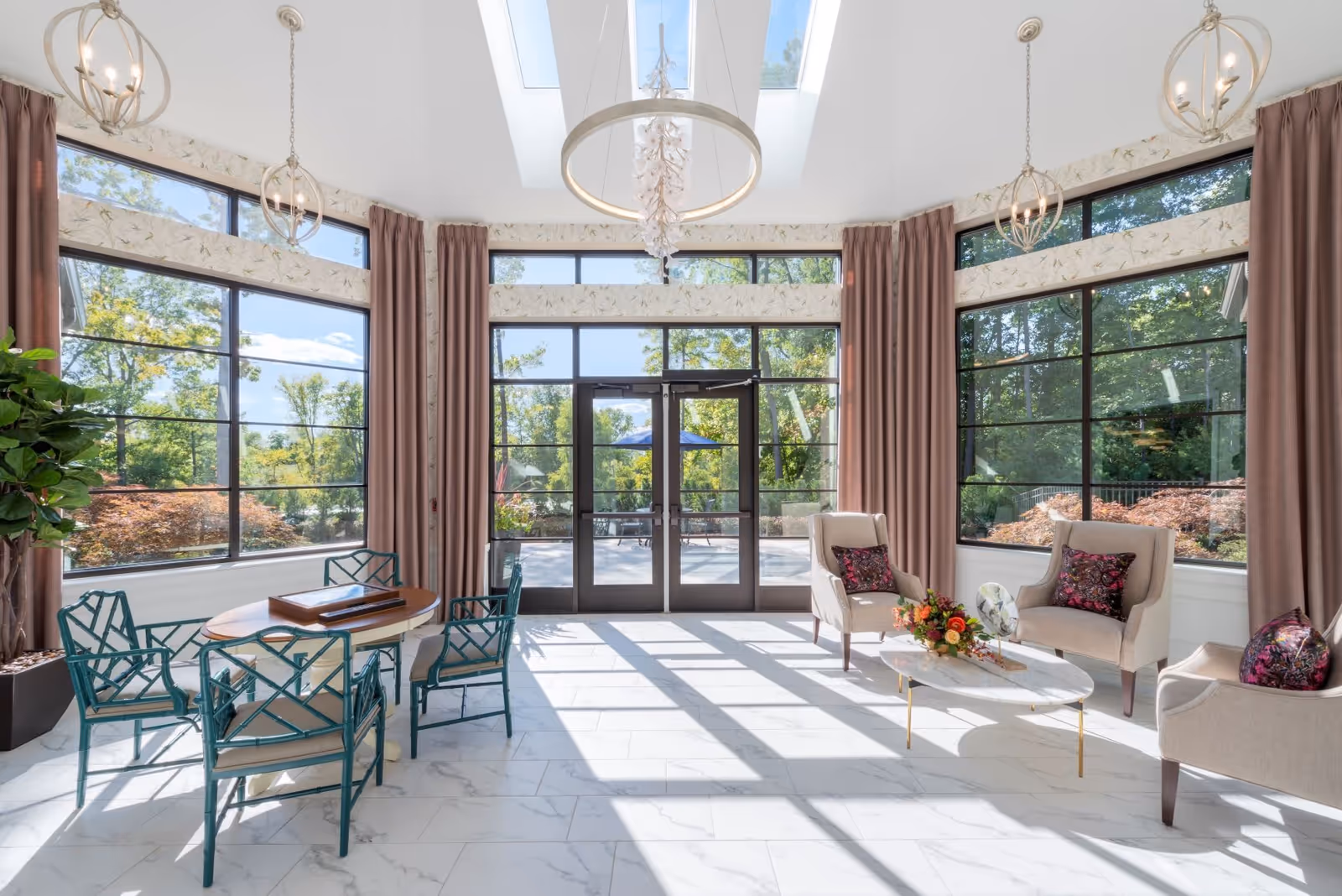 Bright and airy common area with large windows and glass doors leading outside. The room features a round table with four green chairs on the left, and a seating area with three beige armchairs and a white marble coffee table with floral decorations on the right. The space has white marble flooring, tall pink curtains, and modern pendant lights hanging from the ceiling with skylights above.
