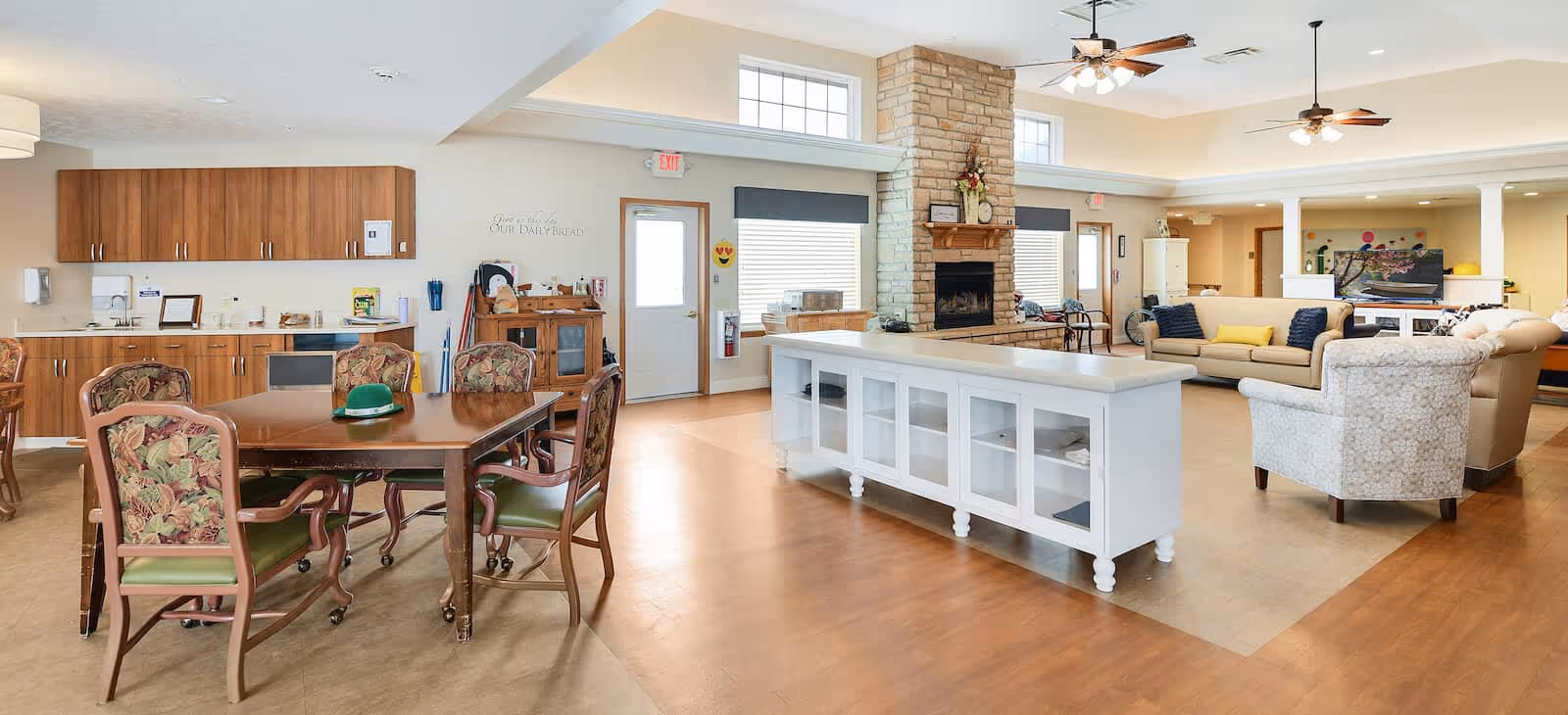 A spacious and well-lit common area in a senior living facility featuring a dining table with six upholstered chairs on the left, a white kitchen island in the center, and a cozy living room area with sofas and armchairs on the right. The room has a stone fireplace, wooden cabinetry, and large windows allowing natural light to fill the space.