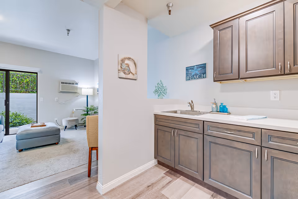 Bright interior view of a kitchenette with dark cabinets and a sink opening to a furnished living area with an ottoman and sliding glass door to a small patio.