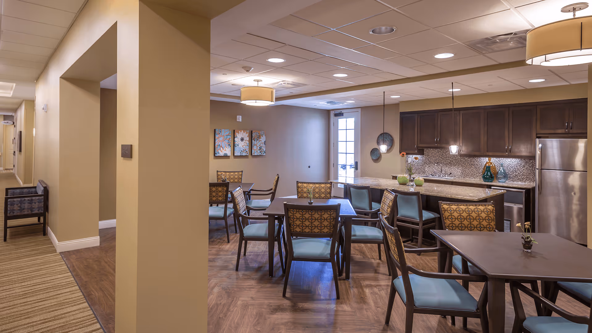 Interior view of a senior living facility common area with multiple tables and chairs arranged for dining or socializing. The room features a kitchen area with dark wood cabinets, a stainless steel refrigerator, and a countertop with bar stools. The walls are painted beige, and there are decorative wall hangings and ceiling lights providing warm illumination.