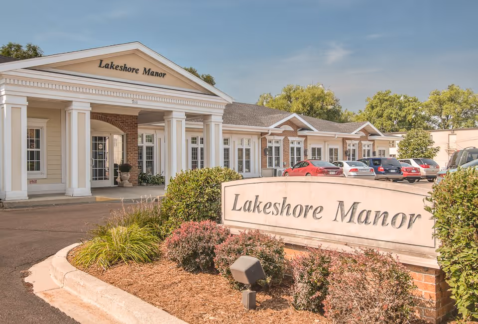 Exterior front view of Lakeshore Manor building with a covered entrance supported by white columns, multiple windows, and a parking lot with several cars. A large sign with the name Lakeshore Manor is visible in the landscaped area near the entrance.