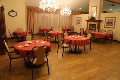 Senior living facility dining room with round tables, red tablecloths, chairs, and chandeliers.