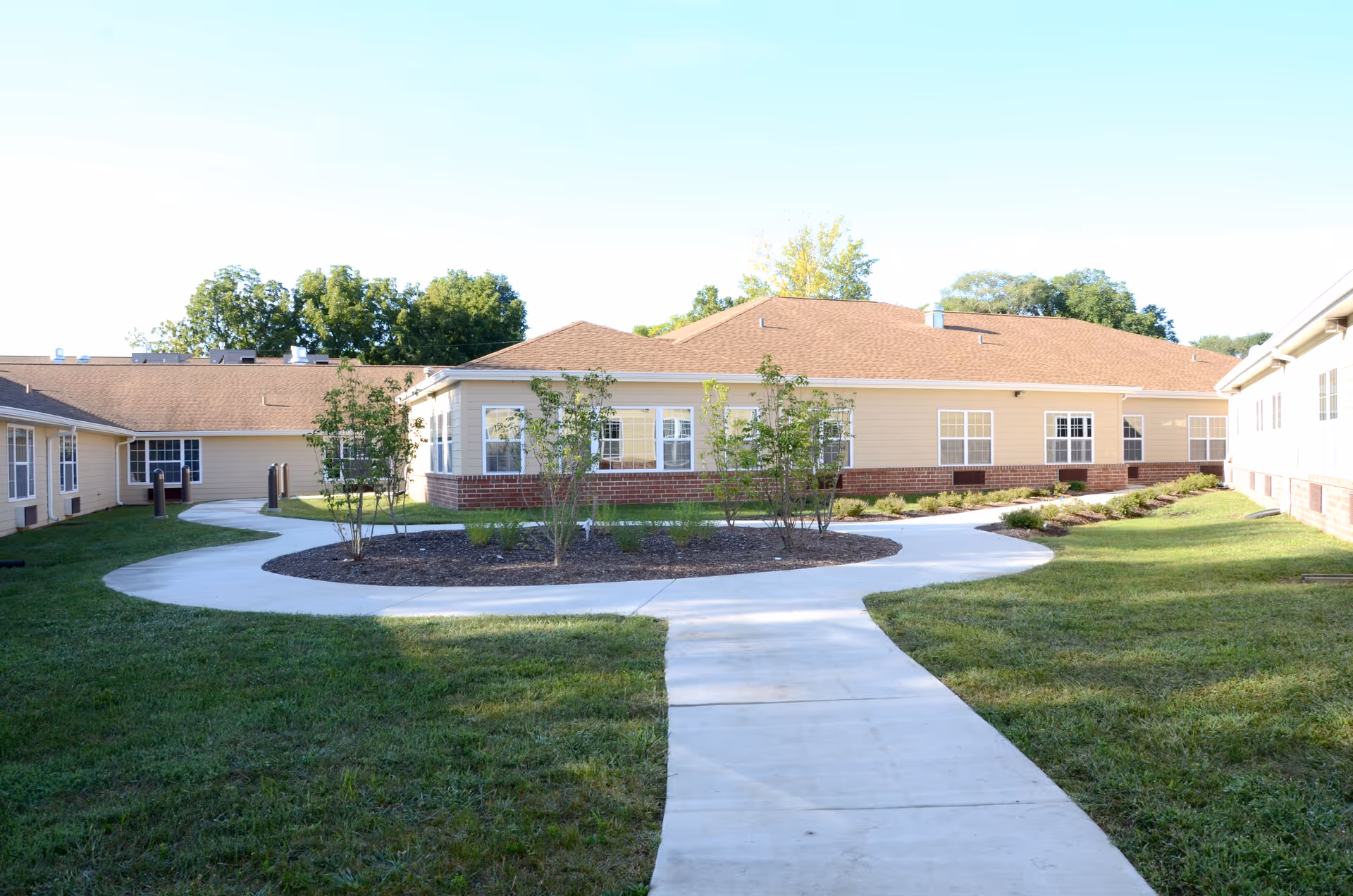 Outdoor view of a senior living facility courtyard with a curved concrete walkway, green grass, small trees, and beige buildings with brown roofs surrounding the area under a clear blue sky.