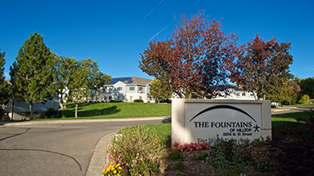 Entrance sign reading 'The Fountains of Hilltop' in front of landscaped grounds and a white senior living building under a blue sky.