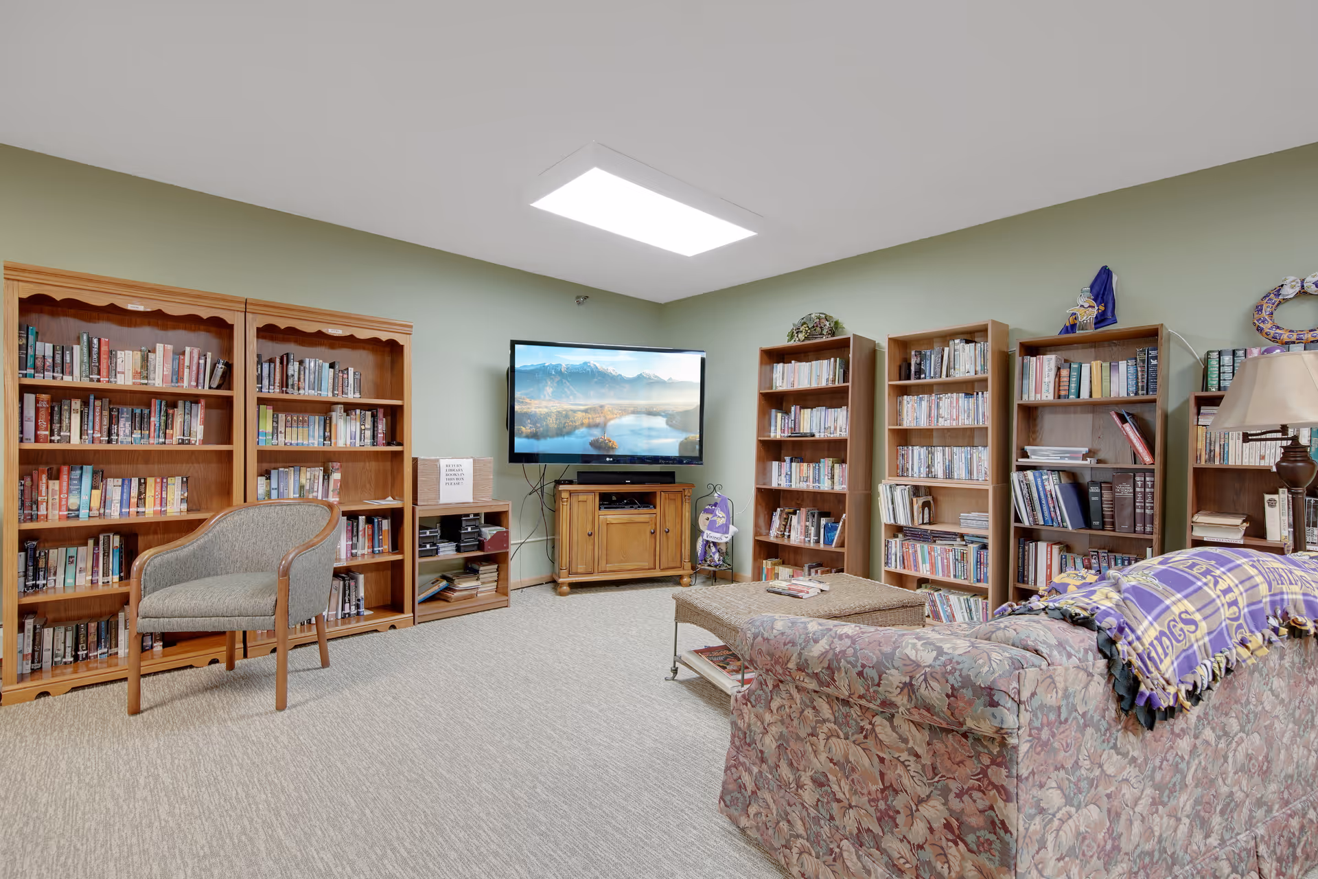 Comfortable common room with multiple wooden bookshelves, a wall-mounted TV, an armchair, coffee table and a floral sofa draped with a blanket.
