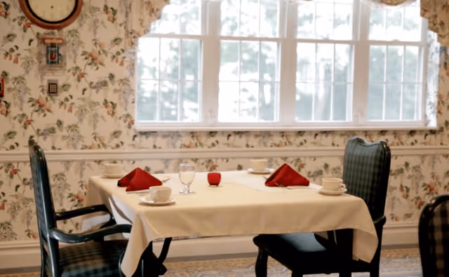 A dining table set for four with a white tablecloth, red folded napkins, cups, saucers, and a glass of water in a room with floral wallpaper and a large window letting in natural light.