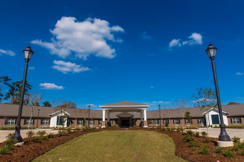 Front exterior view of a single-story healthcare and rehabilitation center building with a covered entrance, surrounded by landscaped gardens and lamp posts under a blue sky with some clouds.