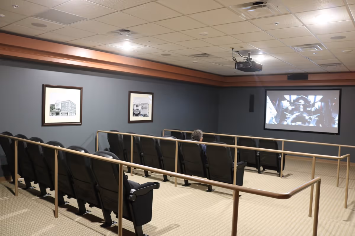 A small movie theater room with rows of black seats and beige railings. A single person is seated watching a movie projected on a screen at the front. The walls are gray with two framed black and white pictures, and the ceiling has recessed lighting and a mounted projector.