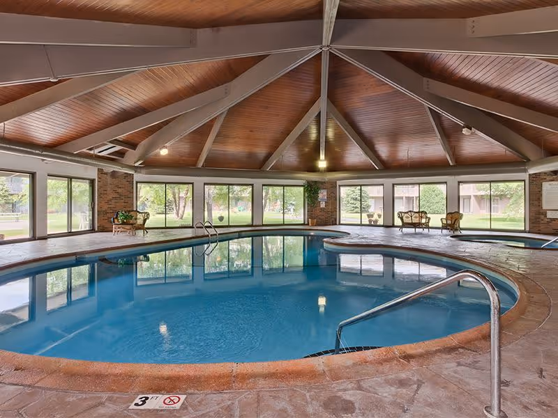 Indoor circular swimming pool in a pavilion-style room with a wooden vaulted ceiling and large windows overlooking the grounds.