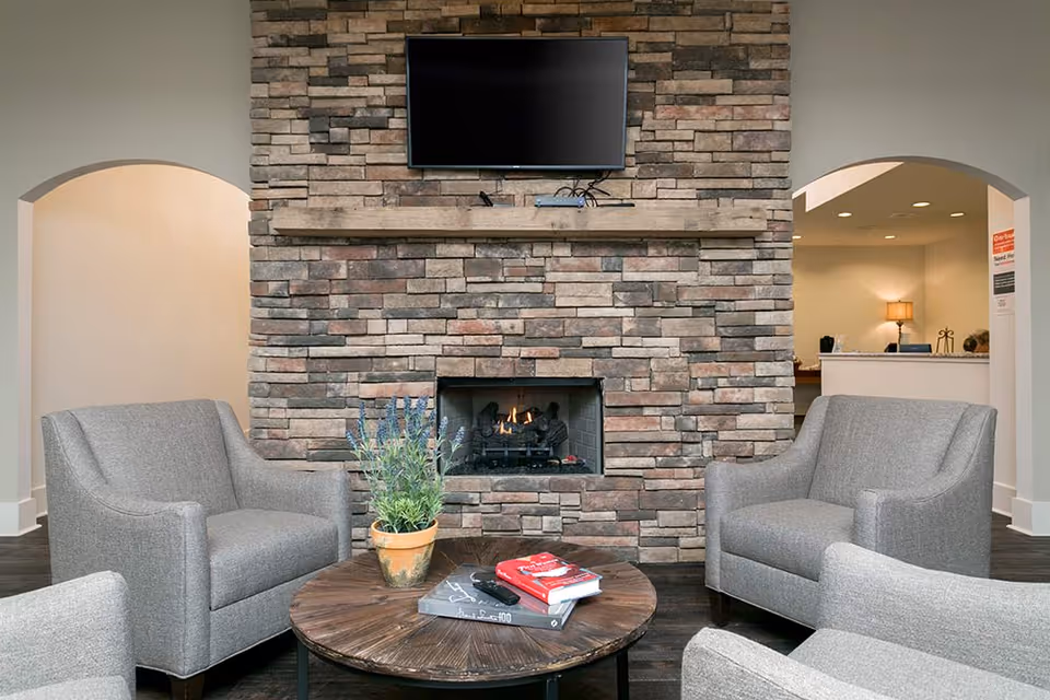A cozy sitting area with four gray upholstered armchairs arranged around a round wooden coffee table. On the table, there is a potted plant and a few books. Behind the seating area is a stone fireplace with a fire burning inside and a flat-screen TV mounted above it. The room has two arched doorways leading to other parts of the facility.