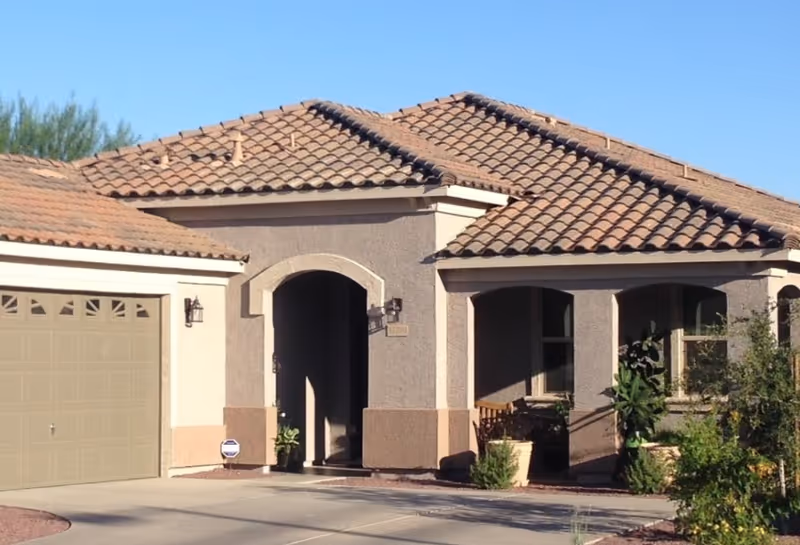 Front exterior view of a single-story house with a tiled roof, an arched entryway, a garage door on the left, and some plants and shrubs near the entrance under a clear blue sky.