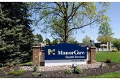 Outdoor view of a sign for ManorCare Health Services, surrounded by landscaping with trees and grass under a clear blue sky.