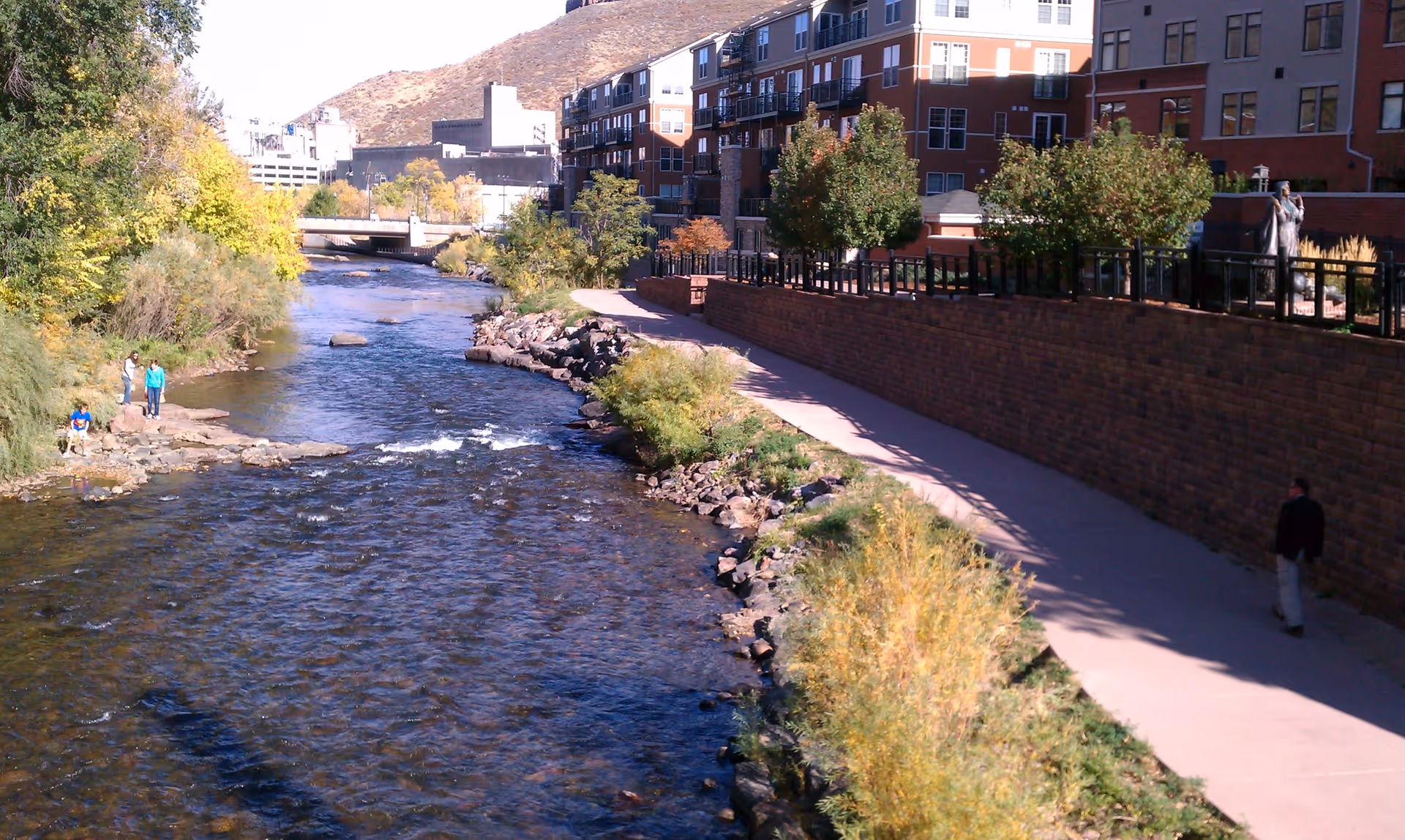 A river flowing alongside a paved walking path with bushes and trees on both sides. On the right side, there is a brick retaining wall with a multi-story residential building behind it. Several people are walking or standing near the river and path. In the background, there is a bridge and a hill.