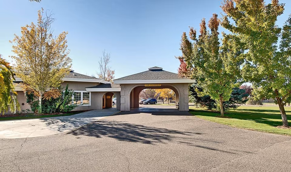 Exterior view of Wellsprings Assisted Living facility entrance with a covered driveway, surrounded by trees with autumn foliage and a clear blue sky.